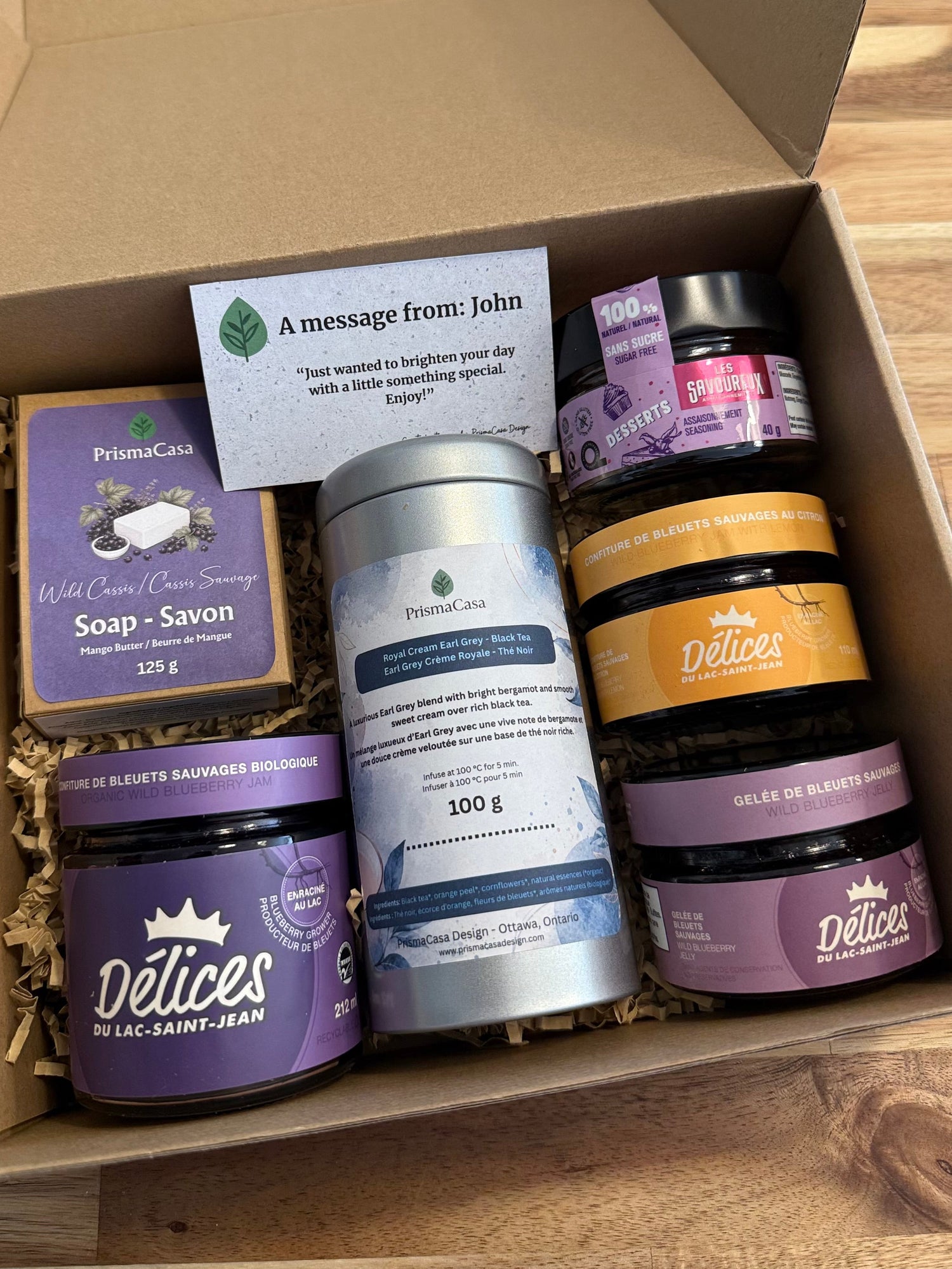 A curated gift box containing various artisanal products including purple and gold jars, a silver tin, and a small card, arranged in a brown cardboard box on a wooden surface.